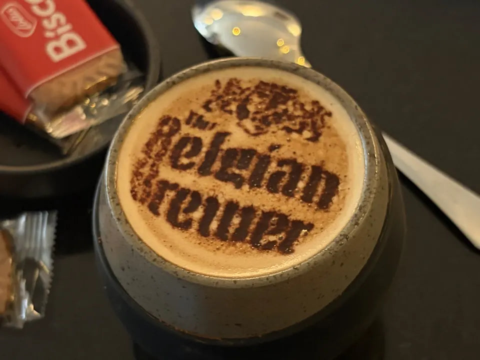 A coffee with The Belgian Brewer crest and name dusted on the foam, Lotus Biscoff biscuits alongside