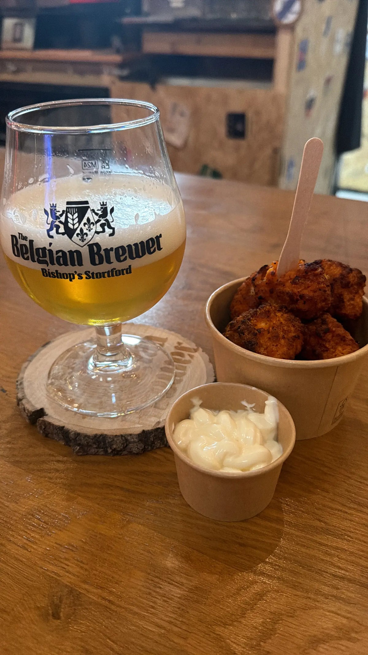 A blond beer in a Belgian Brewer glass beside a pot of fried bites and a dip on a taproom table