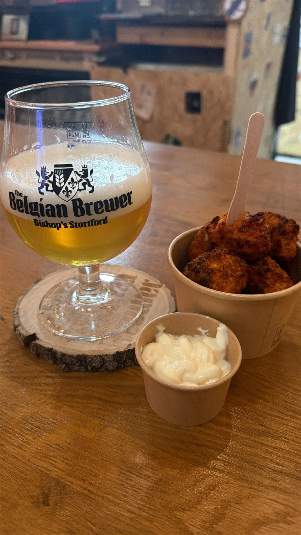 A blond beer in a Belgian Brewer glass beside a pot of fried bites and a dip on a taproom table