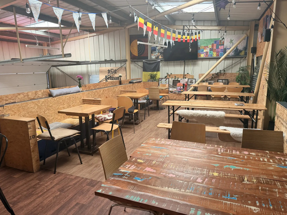 The mezzanine at the Stortford taproom: beer-hall benches and café tables under Belgian flag bunting, with plants, cushions and a Belgian Brewer sign along the far wall