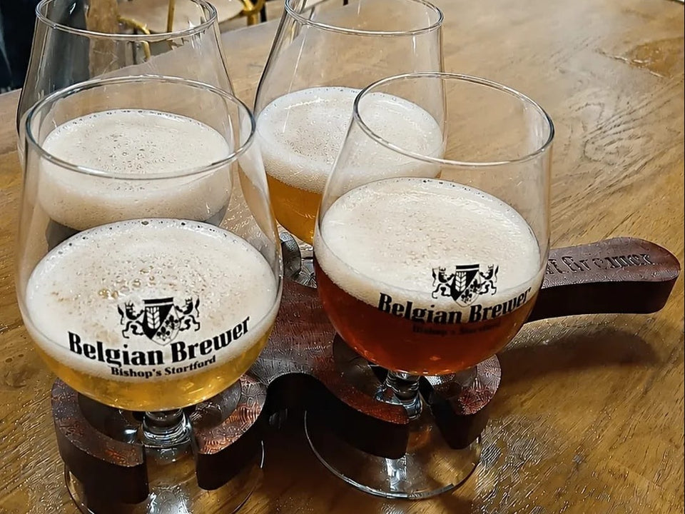A tasting flight of four Belgian Brewer beers, from pale gold to amber, in crested glasses on a wooden paddle